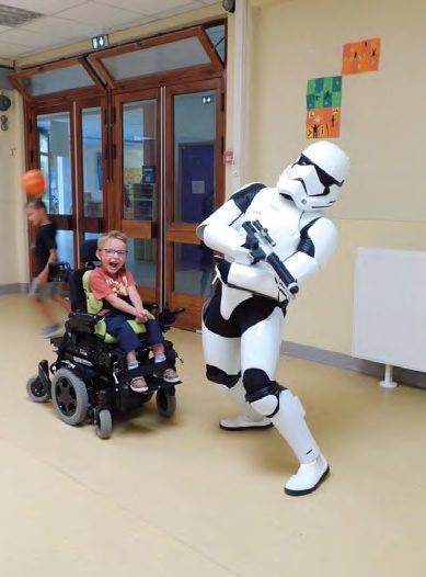 Dans un couloir d'hôpital décoré de dessins, un enfant en fauteuil roulant éclate de rire devant un personnage en armure blanche venu le saluer.