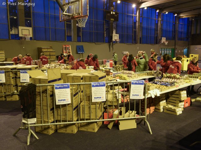 Le gymnase transformé en atelier : des dizaines de tables couvertes de sacs coureurs à garnir, et des bénévoles en veste rouge au fond.