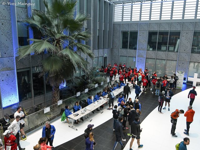 Le hall vitré vu d'en haut : la file des coureurs longe la rangée des tables de retrait.