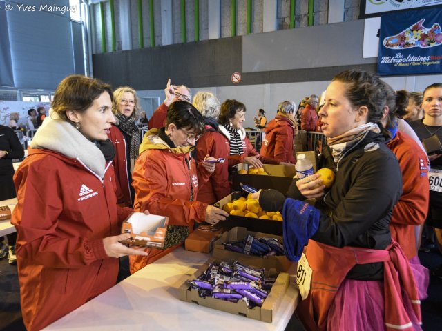 Des bénévoles distribuent oranges et barres à des coureuses encore essoufflées, médaille au cou.